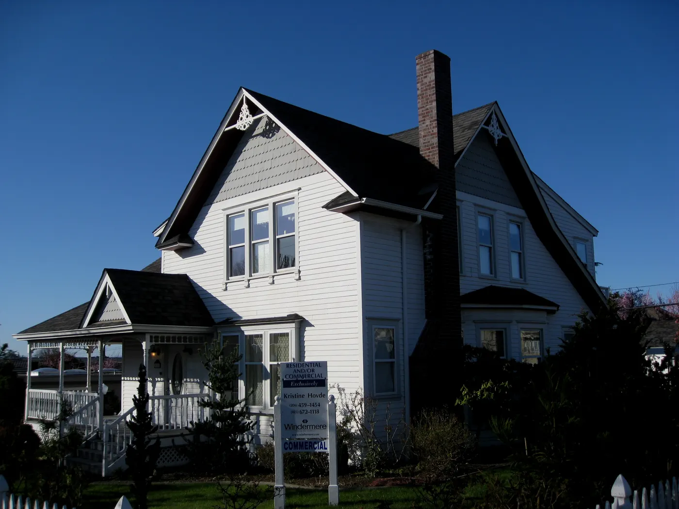 An older two-story Edmonds house with steep gables, a brick chimney, a wraparound porch and shrubs against the foundation