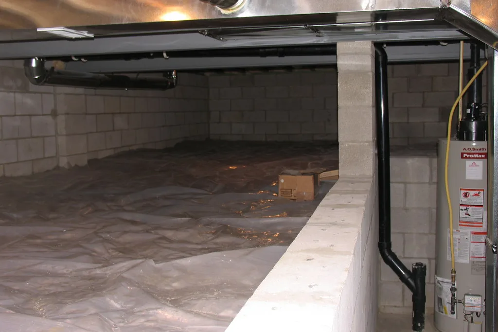 A crawl space with plastic vapor barrier laid over the soil, block walls, a water heater and ductwork overhead