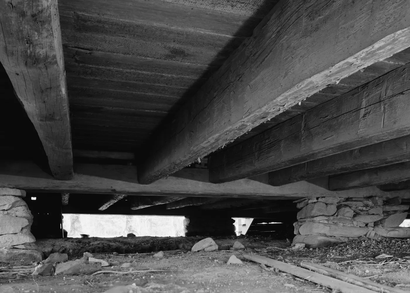 Black-and-white view from inside an old crawl space looking up at heavy timber floor beams resting on stacked stone piers, with debris on the dirt floor