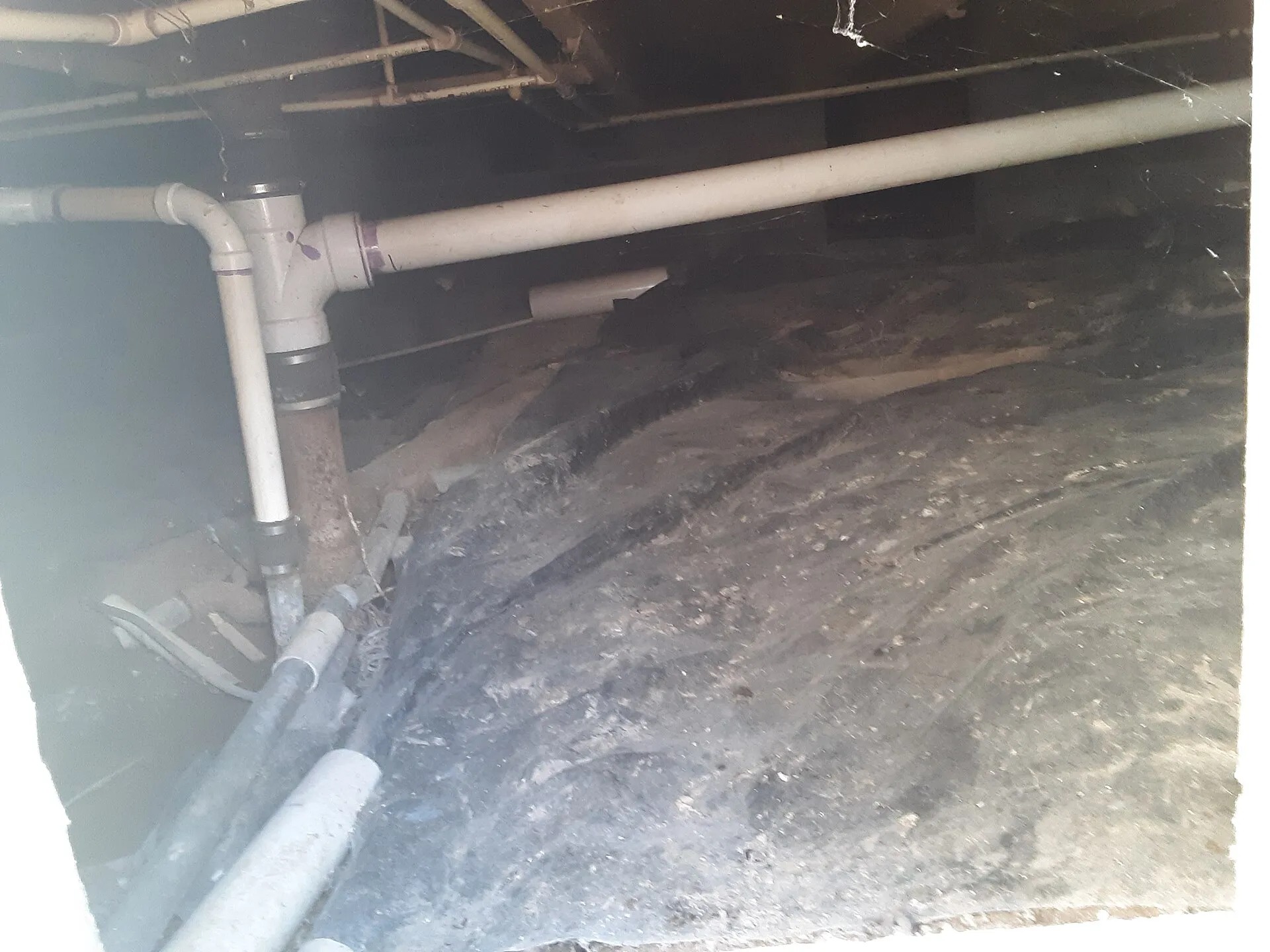A dirty crawl space under a house with bare, debris-covered soil and plumbing running overhead