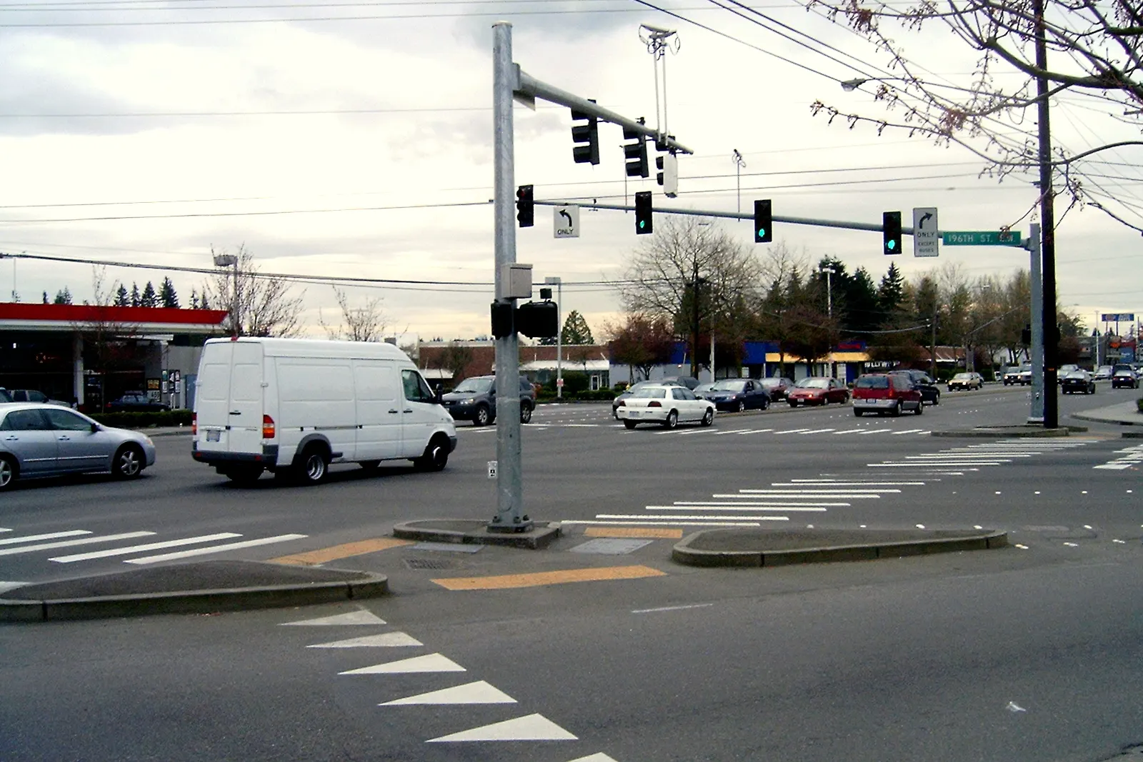 The intersection of 196th Street SW and Highway 99 in Lynnwood, WA, with traffic signals, a gas station and evergreen trees beyond