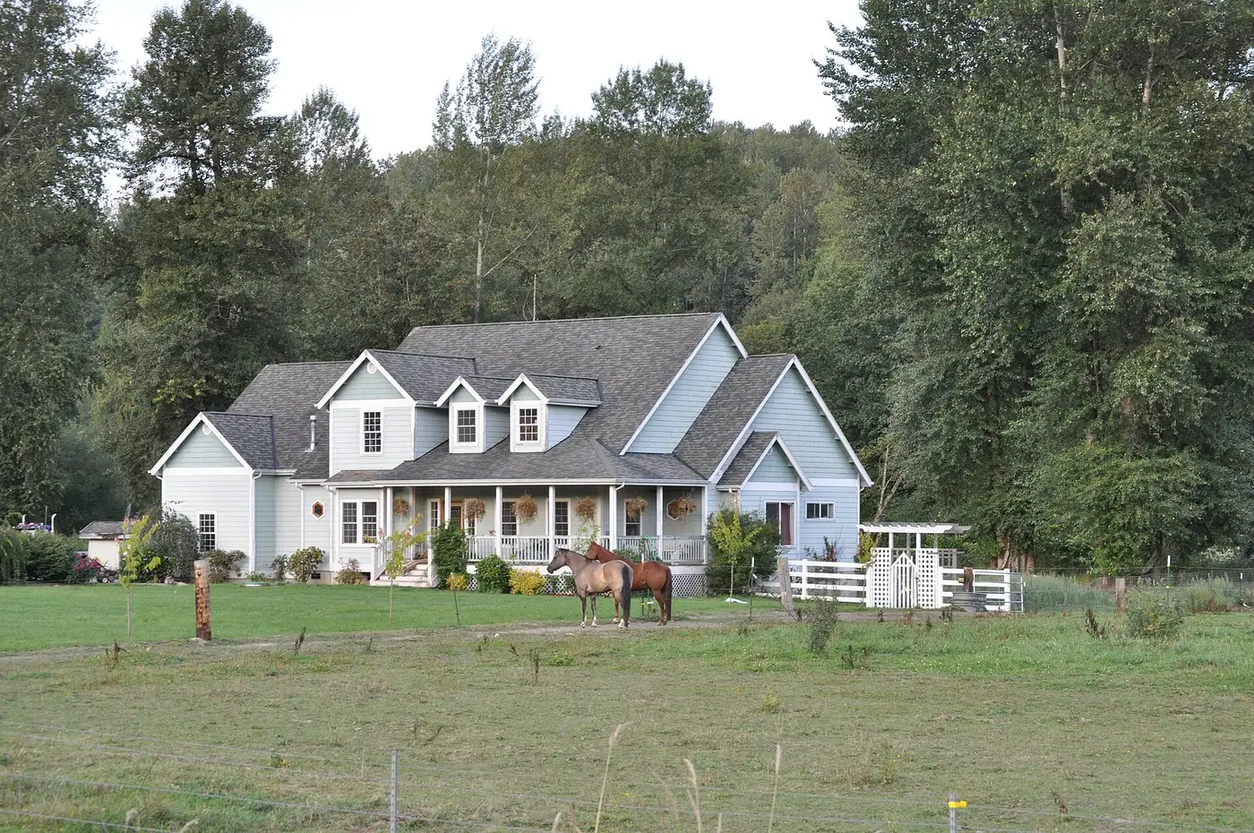 A blue-gray farmhouse with dormers and a wraparound porch, two horses in the pasture in front and a wooded hillside behind, seen from the Centennial Trail near Snohomish, WA