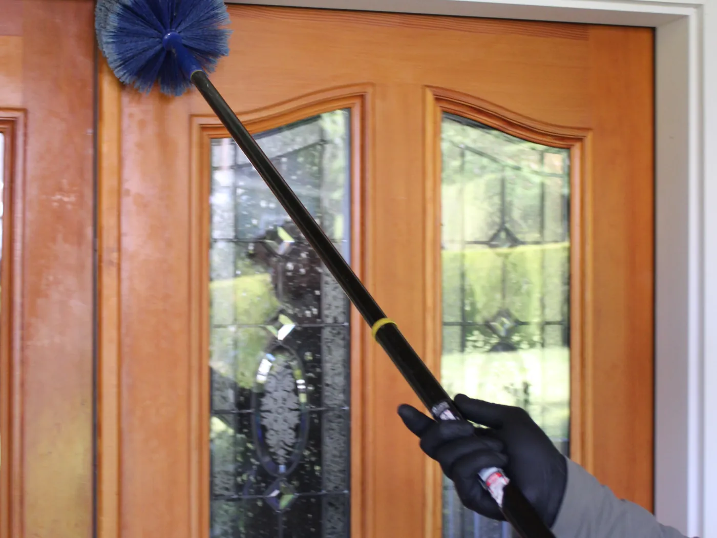 A gloved hand holding an extension web duster up to the top corner of a wooden front door with glass panels