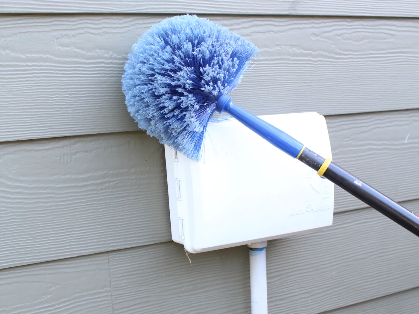 A Pure Guard web duster clearing webs from a utility box mounted on gray lap siding